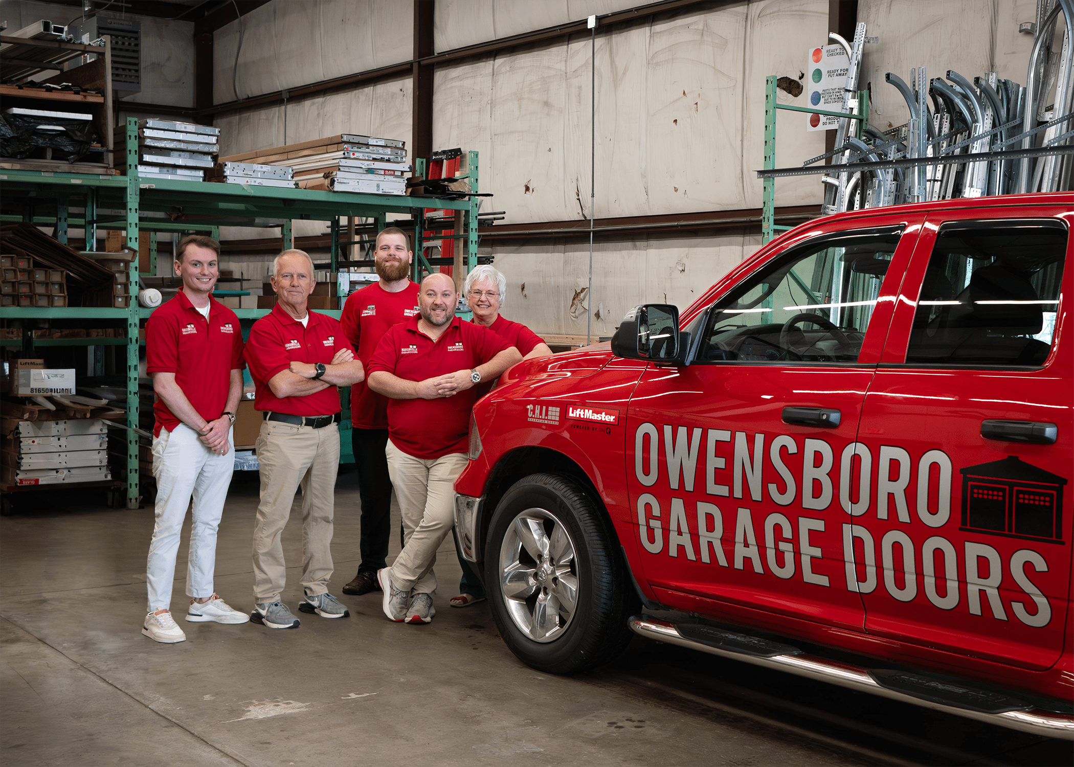 Team Owensboro in matching red shirts stand beside a red truck labeled "Owensboro Garage Doors" in a warehouse, smiling confidently. Shelves are visible in the background.